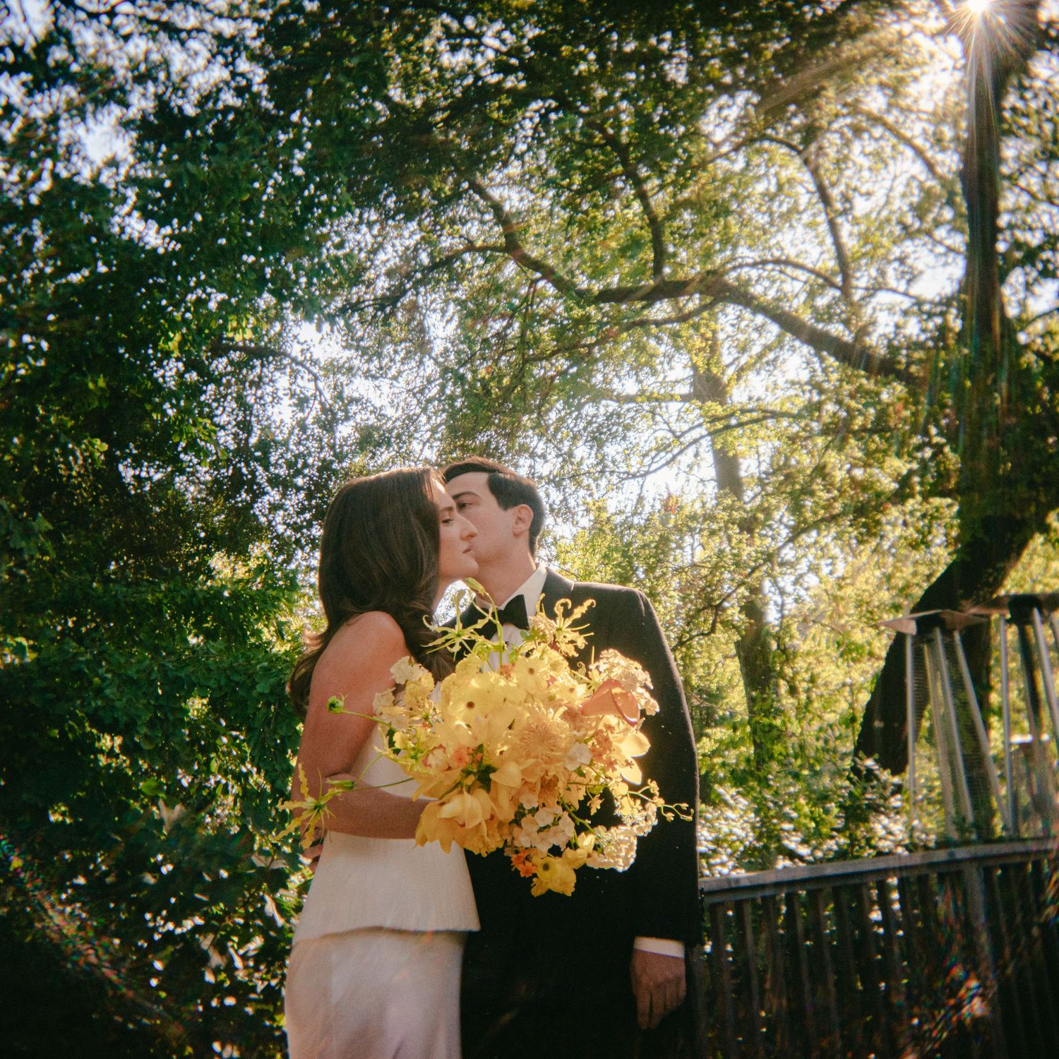 Couple kissing beneath sunlit trees on a wooden deck, the bride holding a yellow and orange bouquet in a romantic wedding portrait.