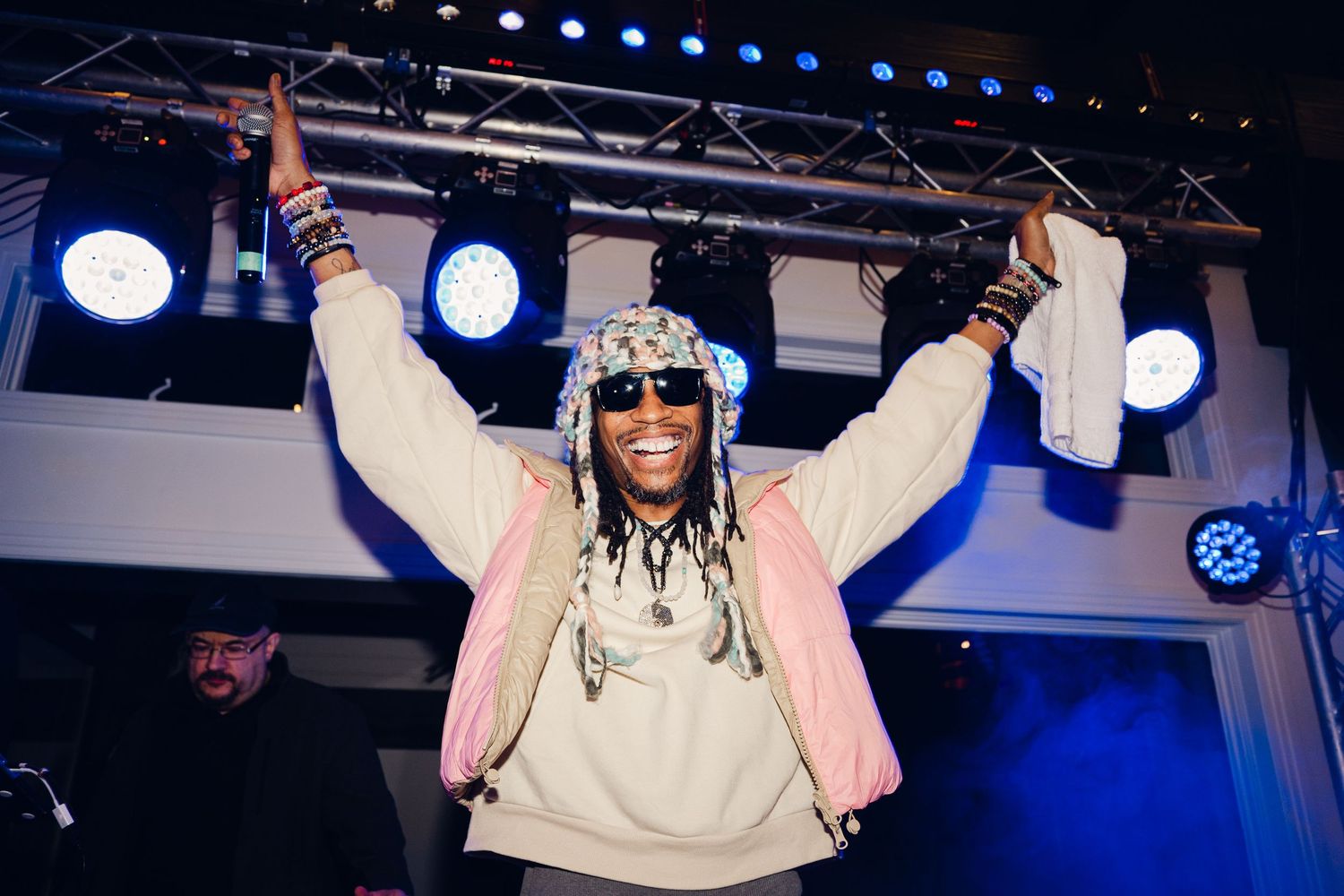 Performer with dreadlocks raises arms triumphantly on stage, wearing colorful bucket hat and hoodie under concert lights.