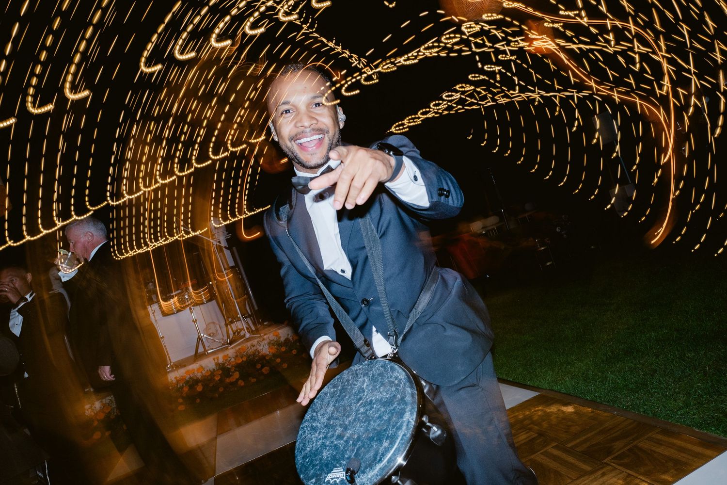 Smiling man in suit pointing at camera surrounded by glowing string lights tunnel at night event.