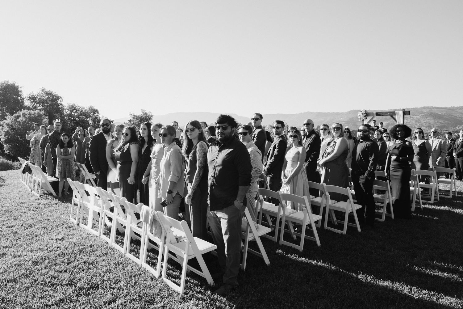 Black and white photo of wedding guests standing at outdoor ceremony with white chairs.