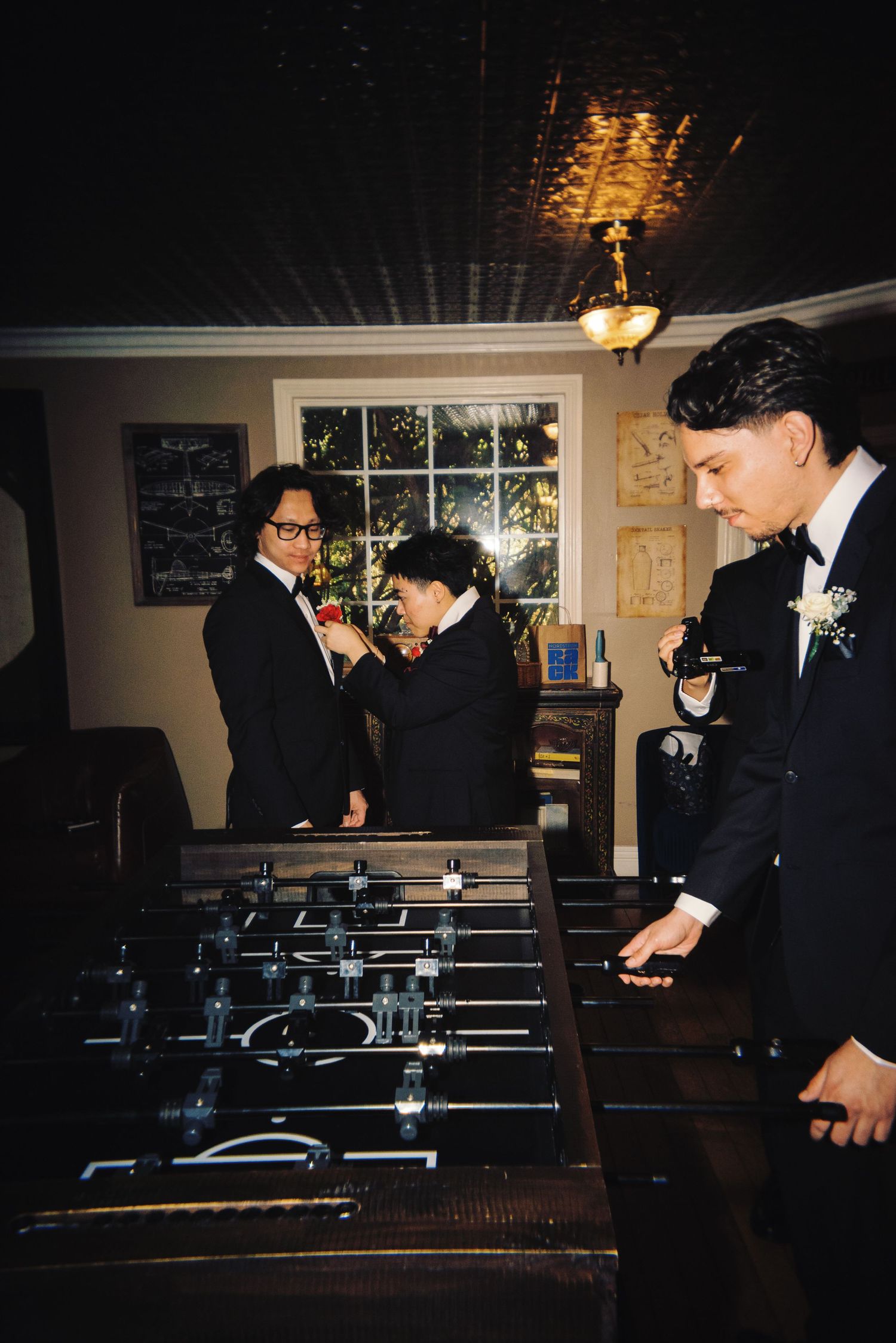 Groomsmen in black tuxedos adjust boutonnieres near a foosball table before the wedding ceremony.