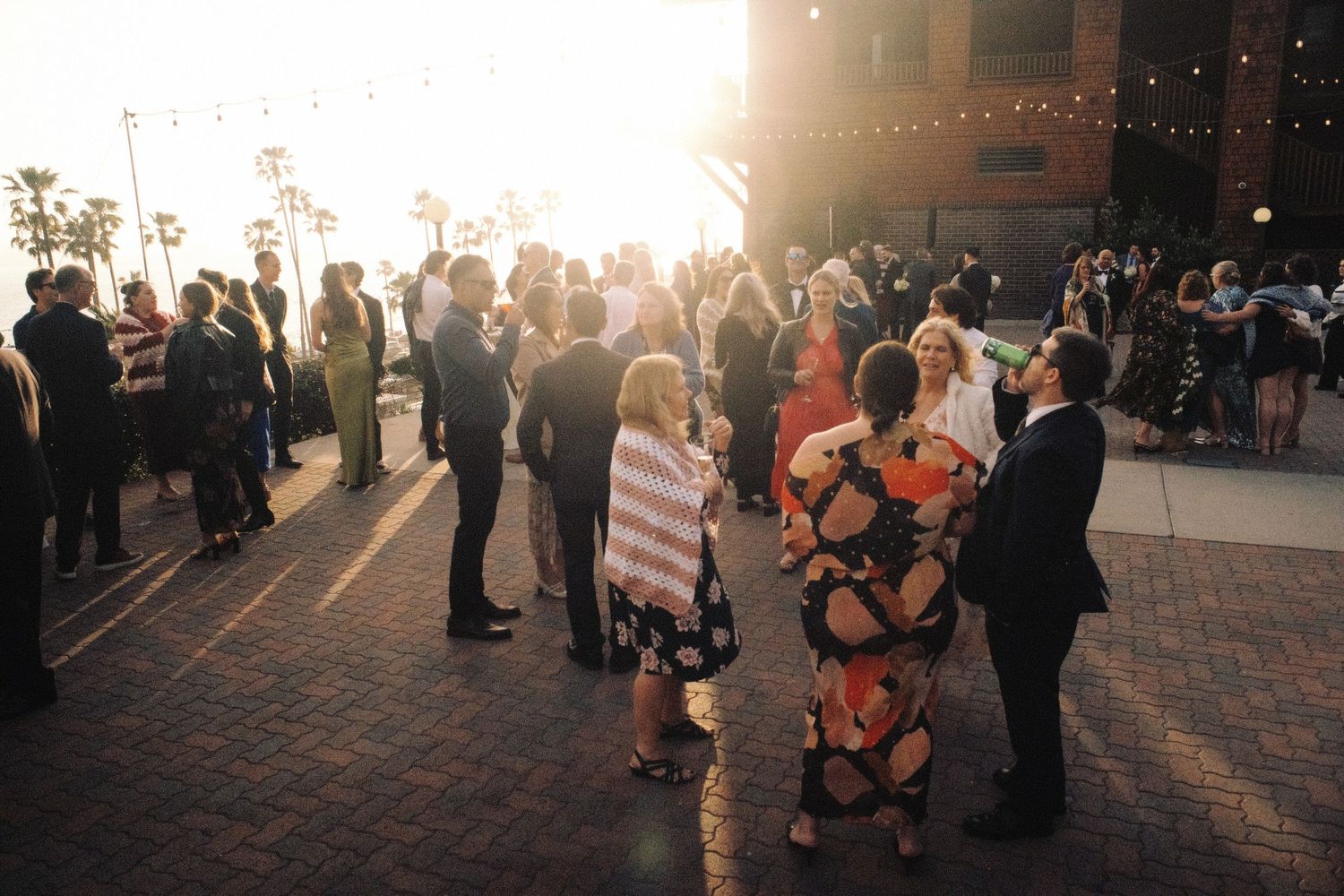 Wedding guests mingle on brick patio in golden sunset light with string lights overhead at outdoor venue.