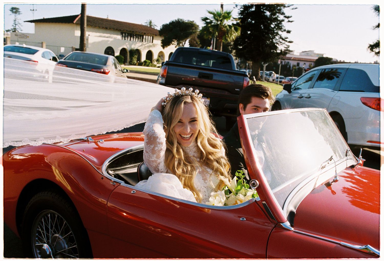 Smiling bride in lace gown and floral crown rides in vintage red convertible with groom after wedding.