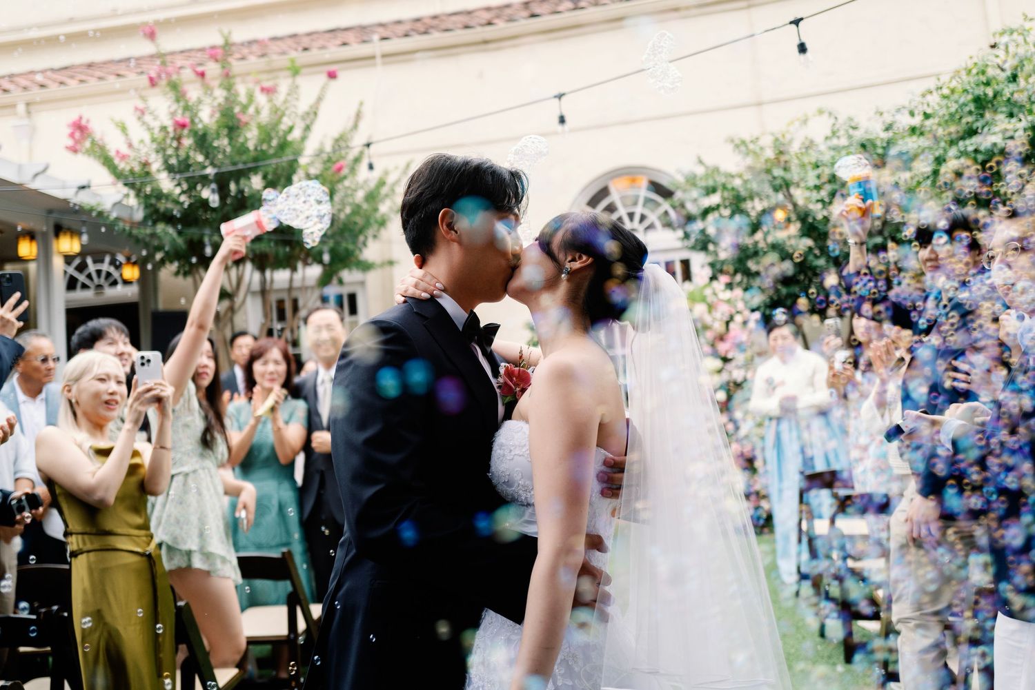 Newlyweds share first kiss surrounded by colorful confetti and cheering guests at outdoor wedding.