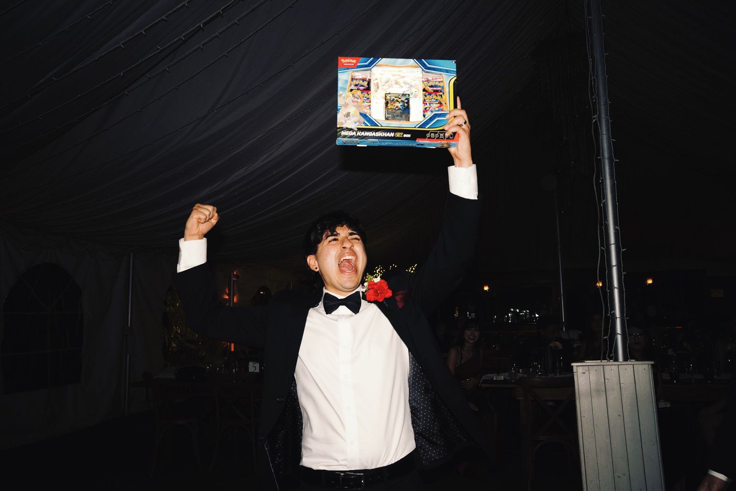 Groom in tuxedo triumphantly holds up a Pokemon card gift box at a wedding reception under a dark tent.
