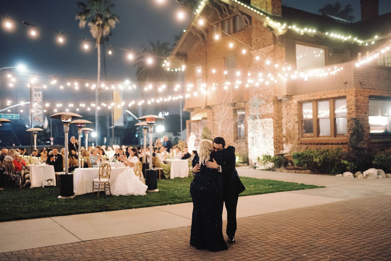 Couple shares first dance outdoors under string lights at elegant night wedding reception with guests seated nearby.