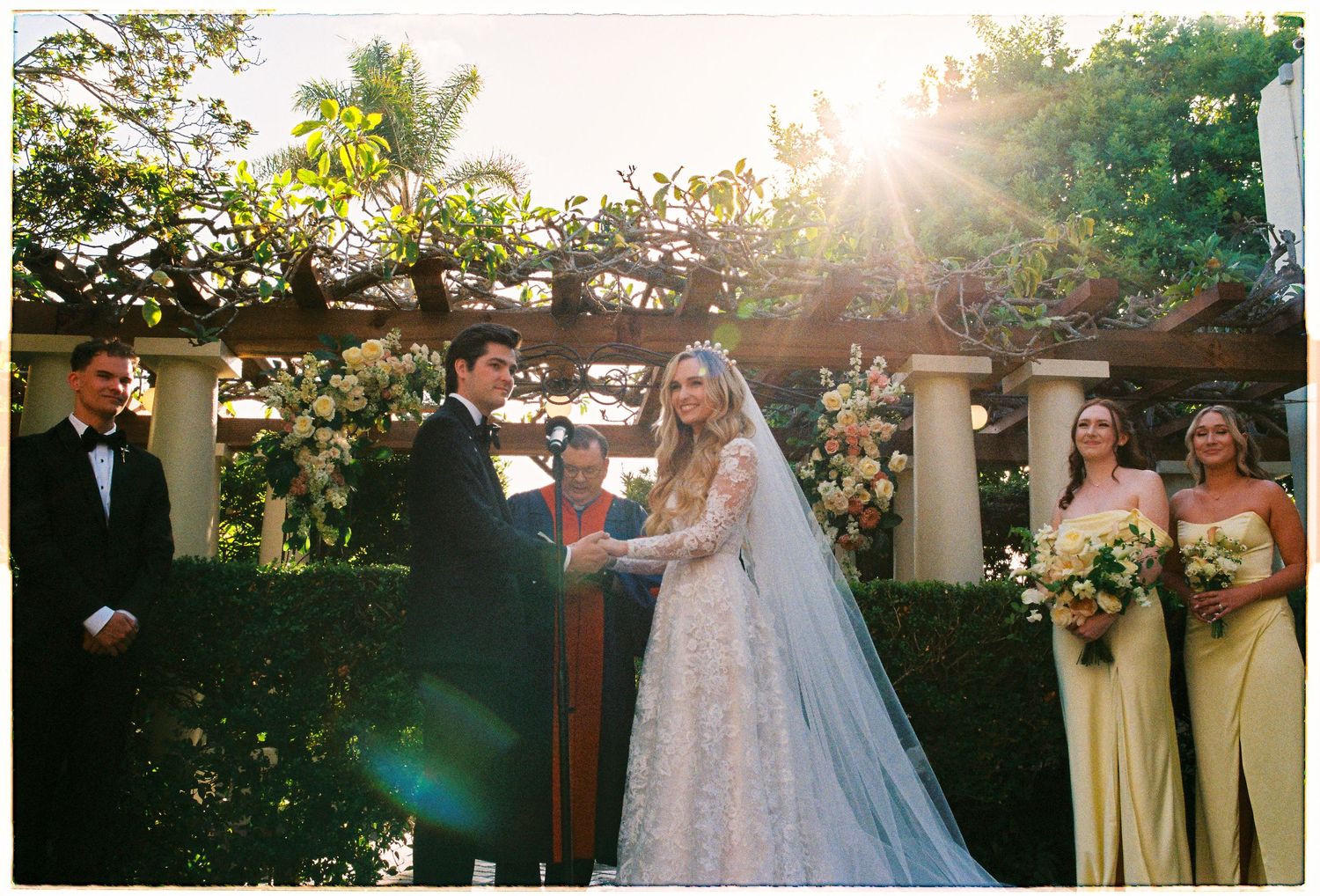 Bride and groom exchange vows under floral pergola with bridesmaids in yellow dresses at sunset.