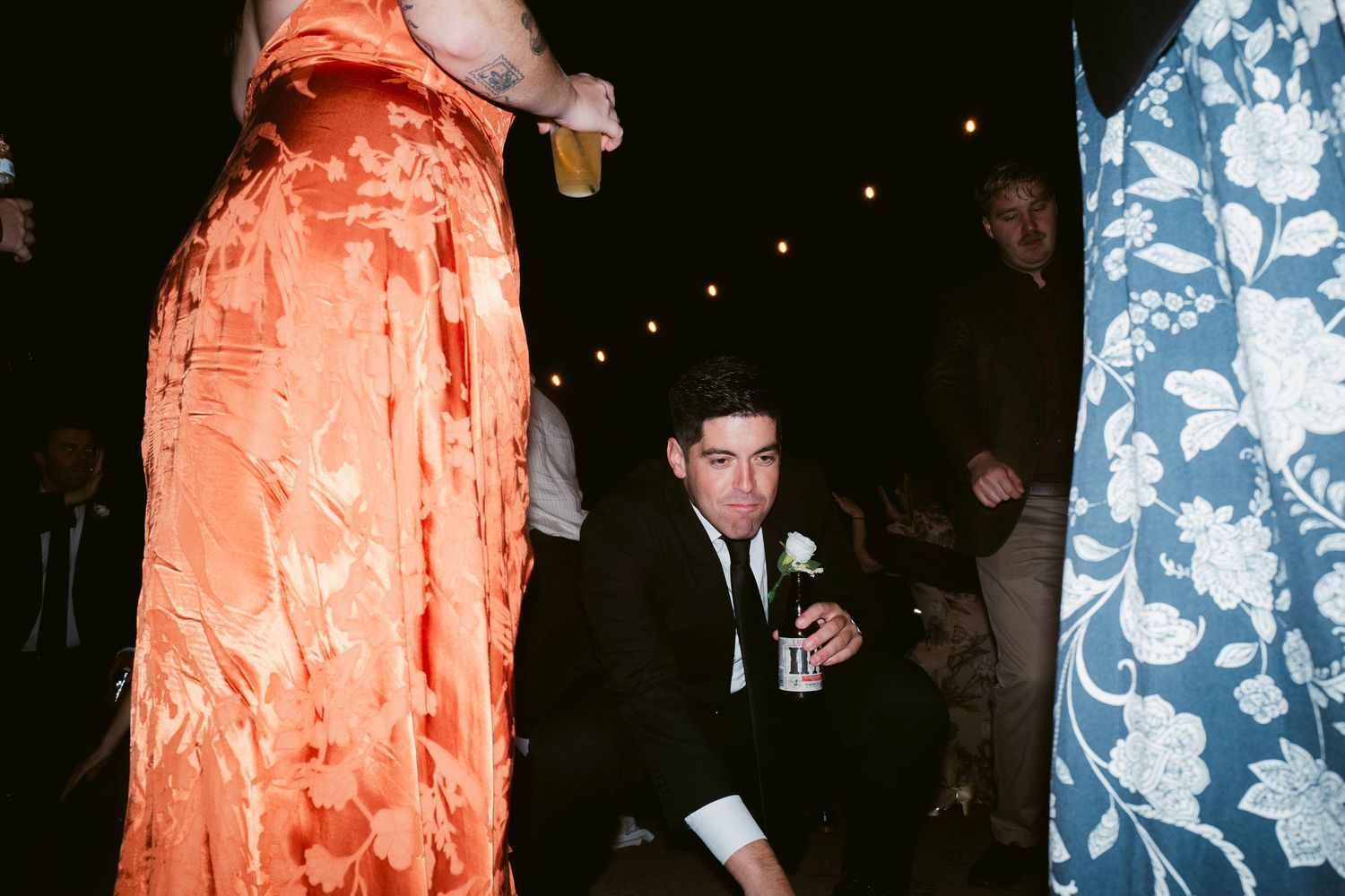 Young man in black suit crouching at colorful wedding reception surrounded by guests in bright attire.