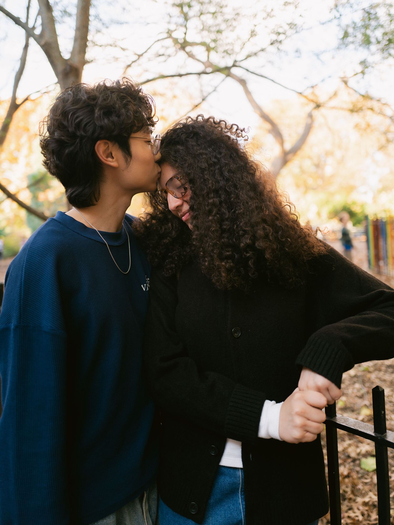 Young man tenderly kissing his girlfriend on the cheek near a park fence in autumn.