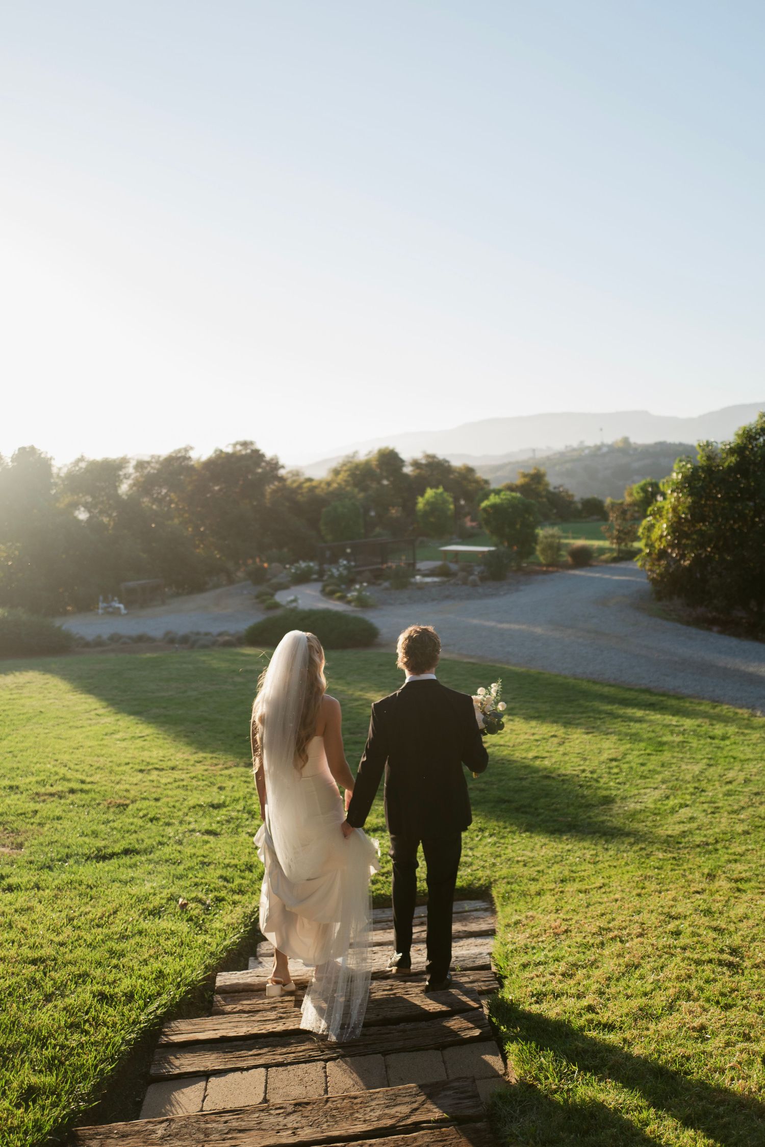 Bride and groom walk hand-in-hand down a stone path through lush green hills at golden hour.