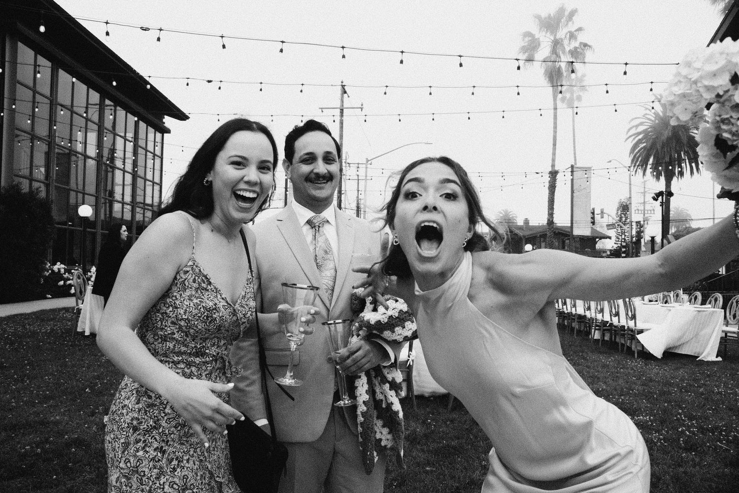 Black and white photo of bride excitedly posing with guests at outdoor wedding reception with string lights.