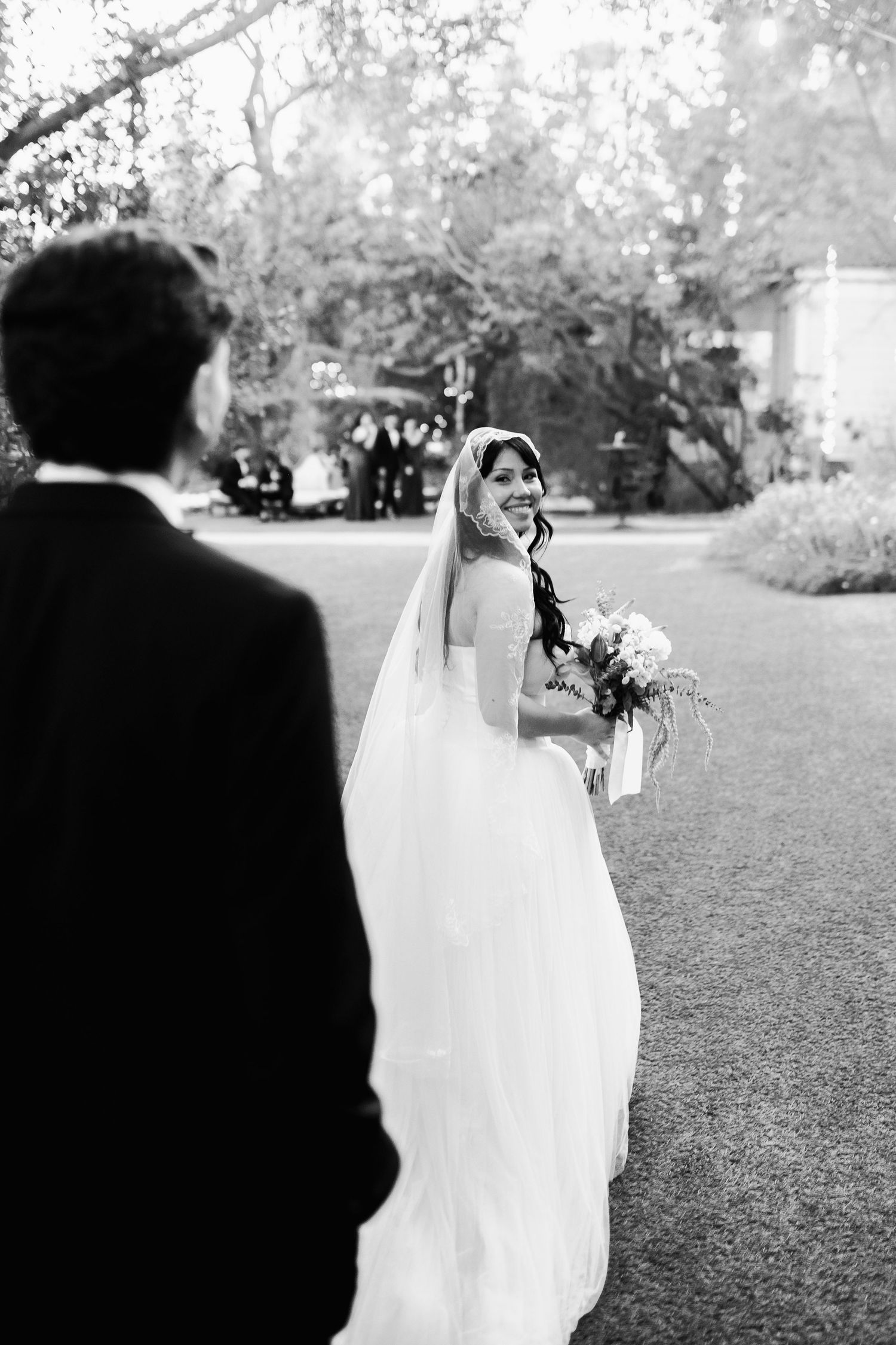 Black and white photo of smiling bride in veil looking back at groom during outdoor shoot.
