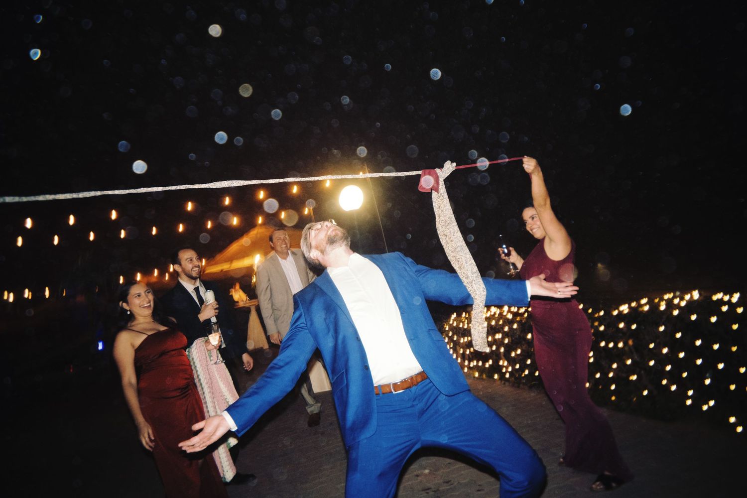 Man in blue suit dramatically dancing at an outdoor wedding reception under glowing string lights.