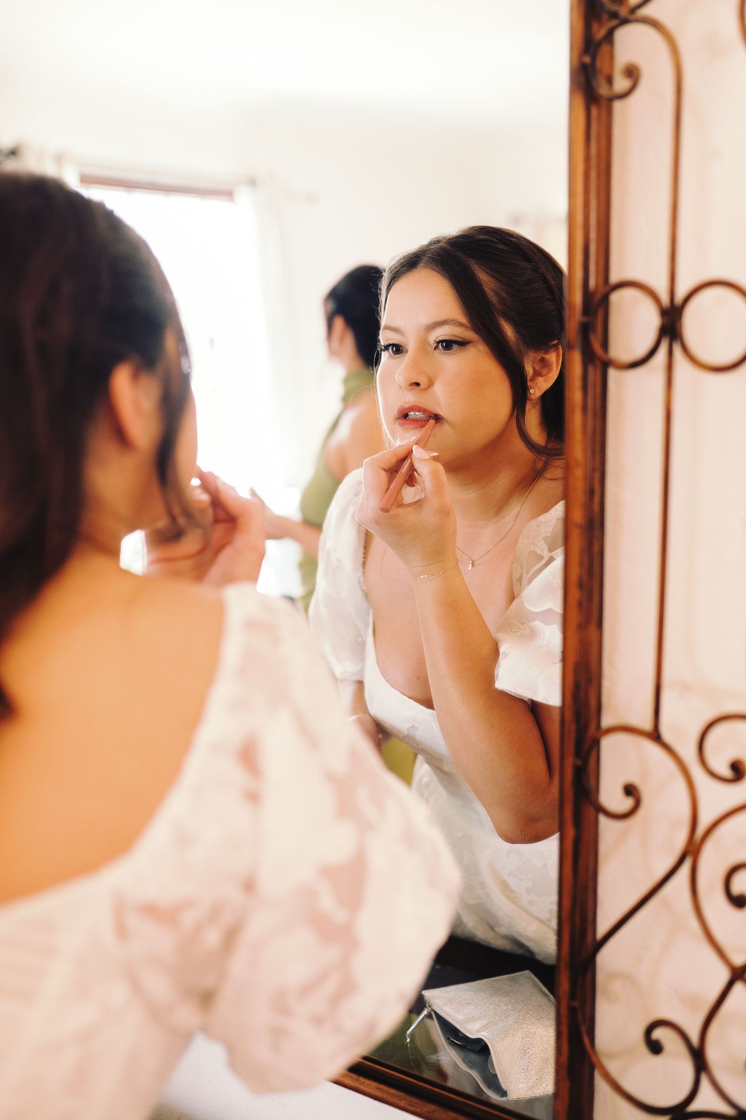 Bride applies lipstick while gazing into ornate mirror during elegant wedding day preparations.