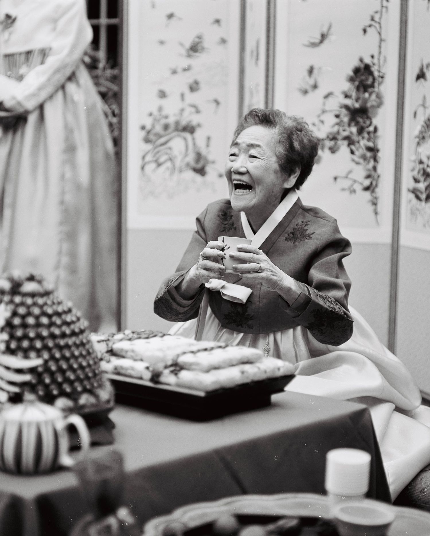 Elderly Korean woman in hanbok laughing joyfully at traditional wedding celebration table.