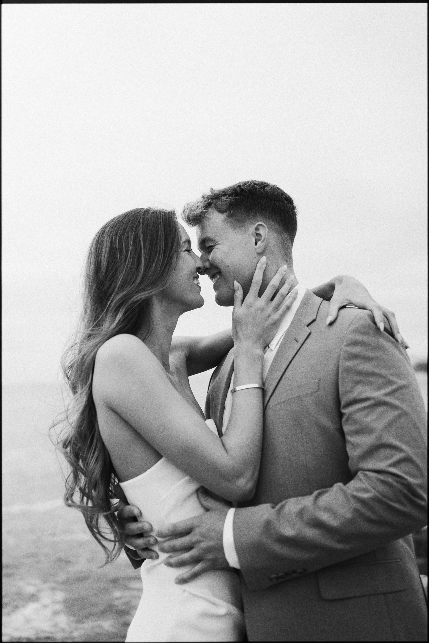 Black and white romantic photo of an engaged couple nearly kissing by the ocean.