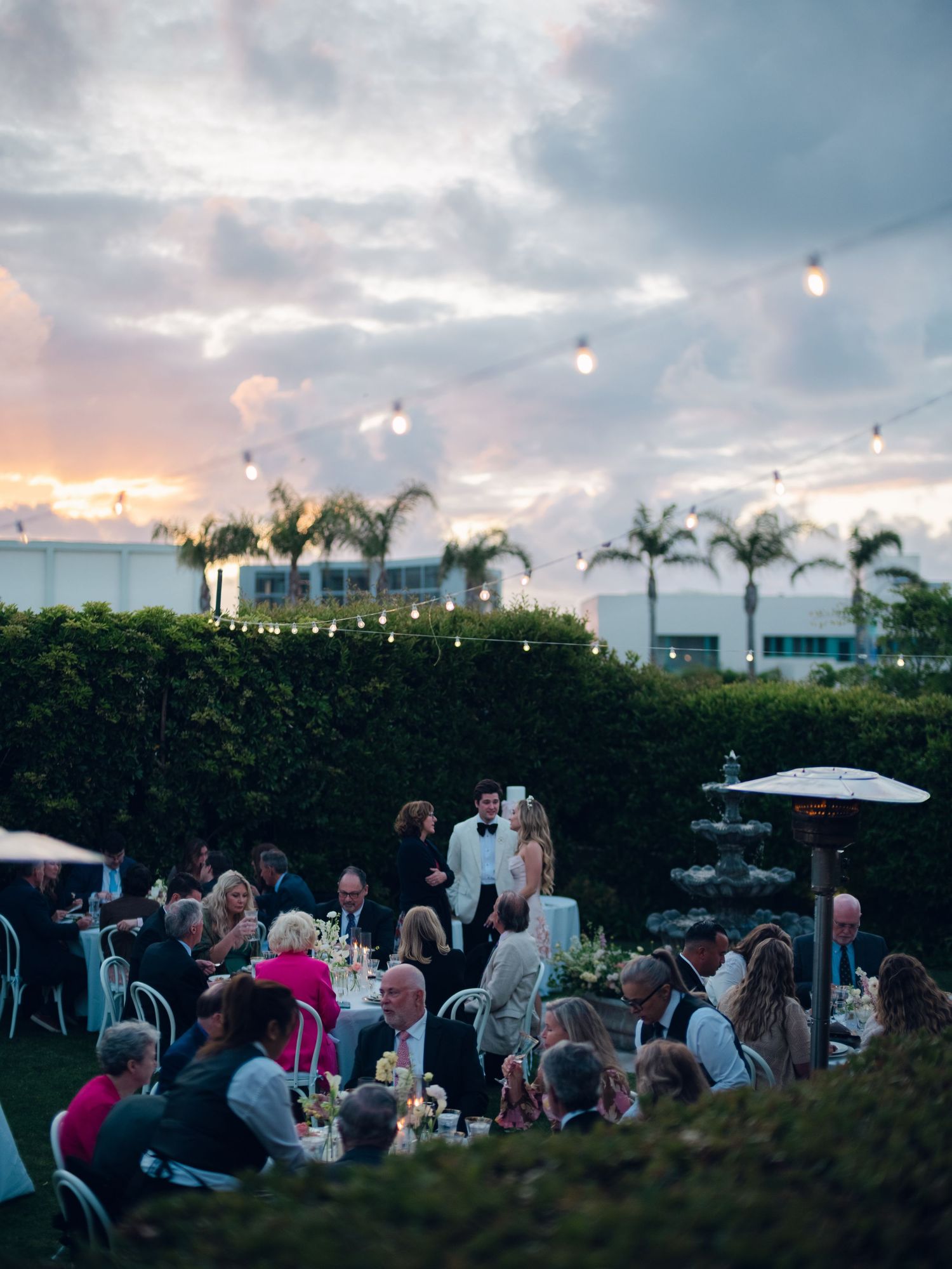 Aerial view of an outdoor wedding reception at sunset with string lights, palm trees, and elegantly set tables.