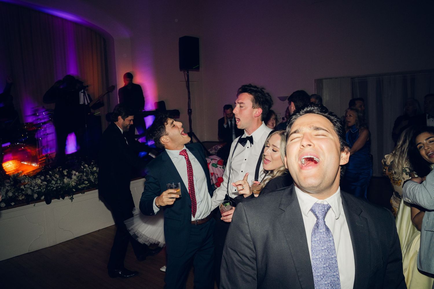 Wedding guests laugh and sing along to music on the dance floor under purple ambient lighting.