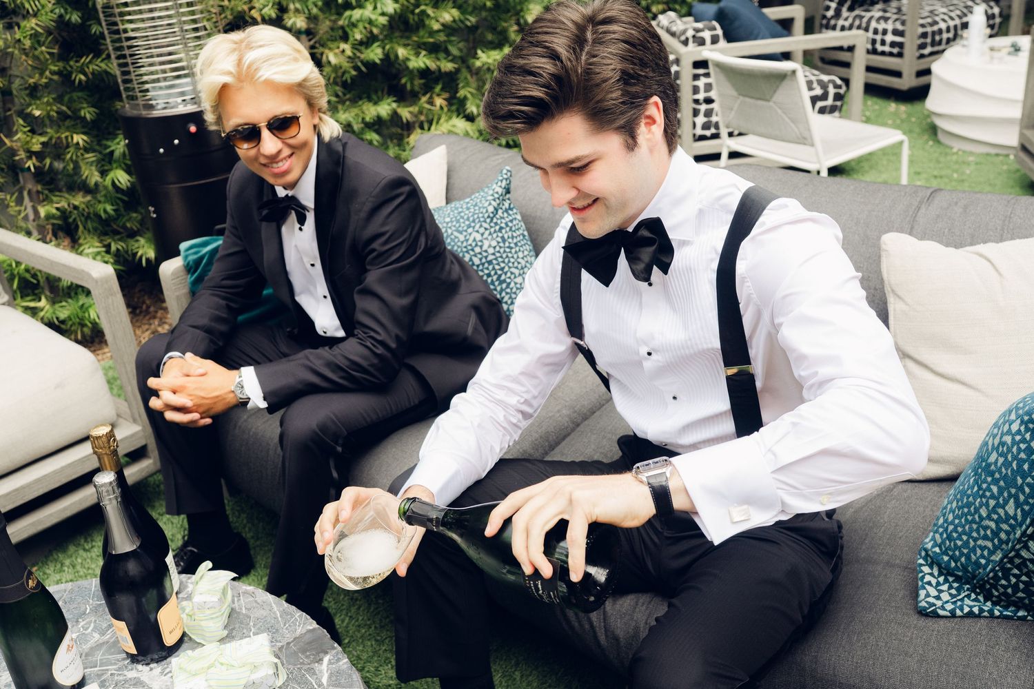 Two men in black tuxedos relaxing outdoors at wedding, one pouring champagne while seated on gray patio furniture.