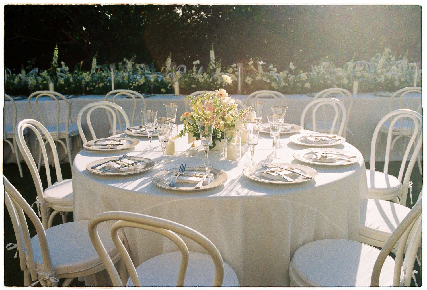 Outdoor wedding reception with round white tables, bentwood chairs, and soft floral centerpieces in garden setting.