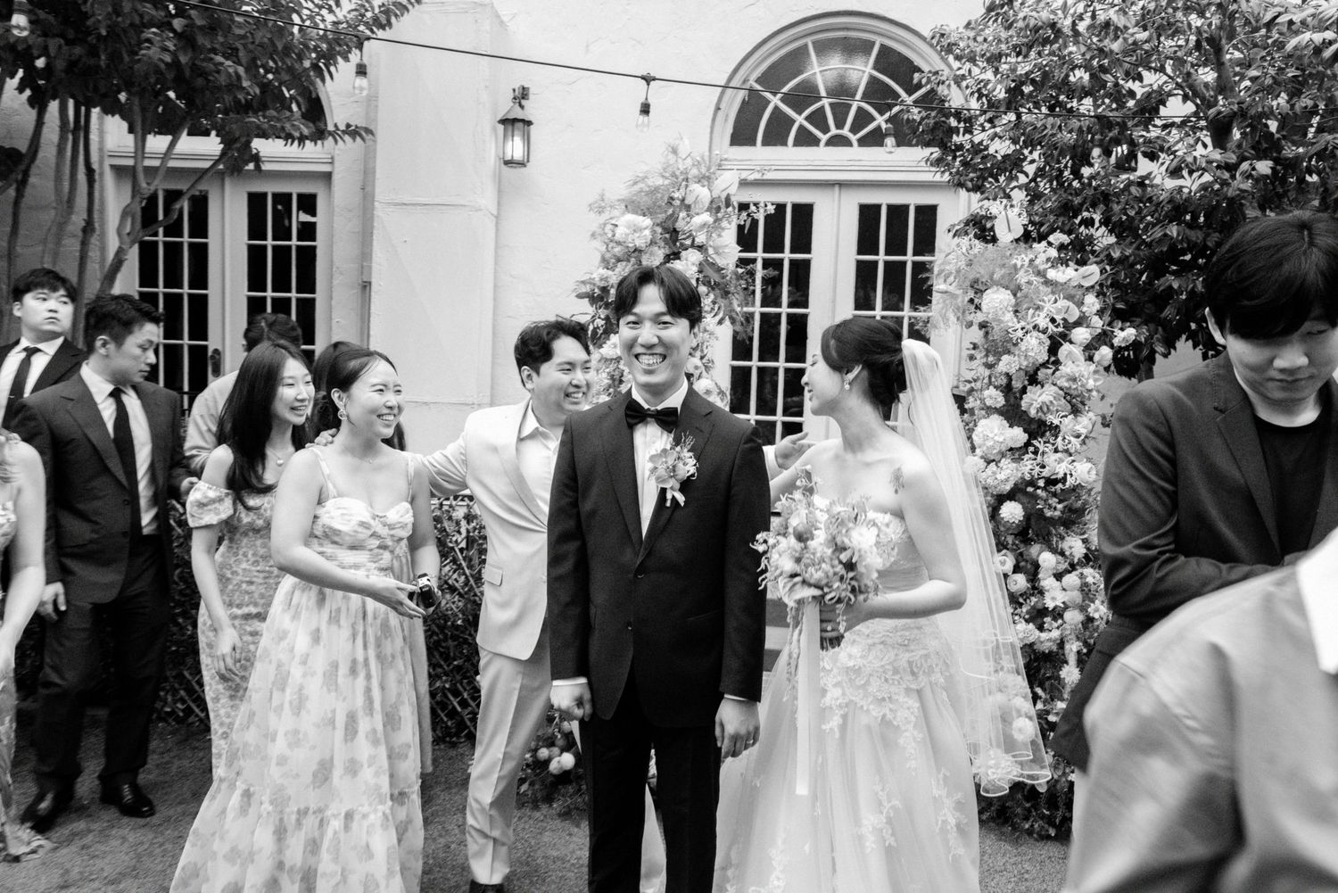 Black and white photo of wedding couple walking with smiling guests outside floral decorated venue.