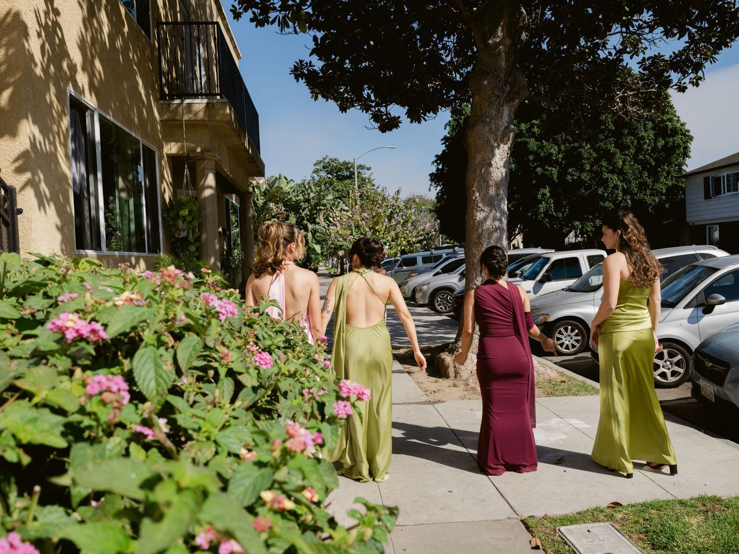 Bridesmaids in olive green and burgundy satin gowns walking along a sunny sidewalk lined with pink flowers.
