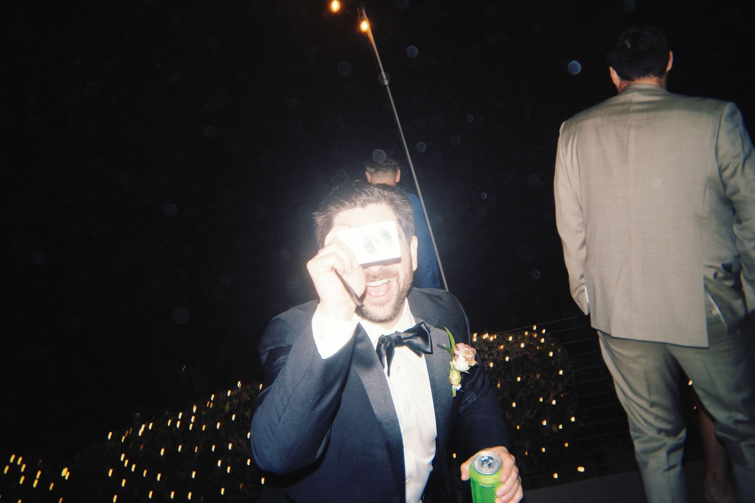 Man in tuxedo with bow tie covering eyes with hands at outdoor evening wedding celebration with sparklers.