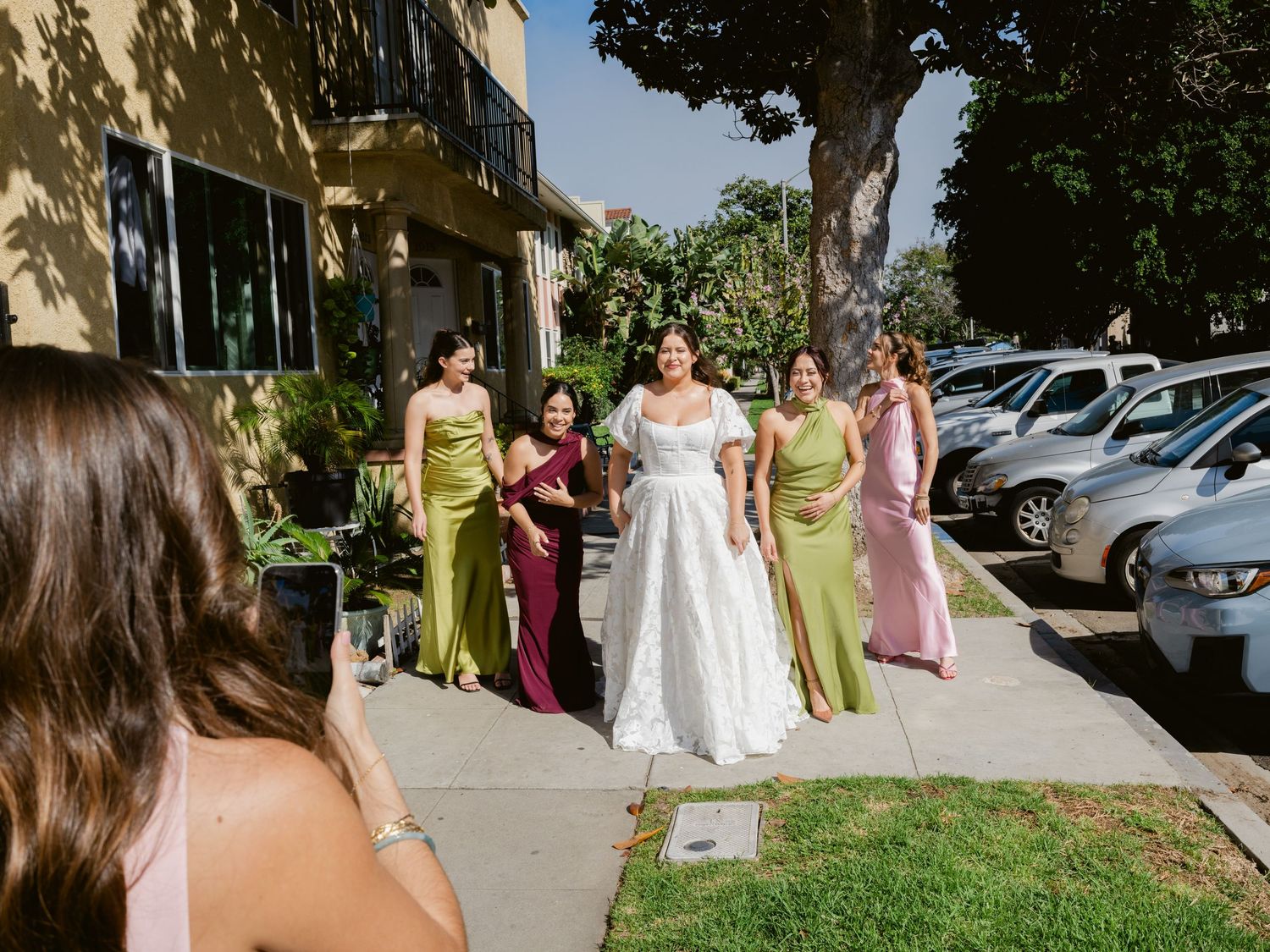 Bride in white lace gown poses with bridesmaids in colorful satin dresses on a sunny suburban sidewalk.