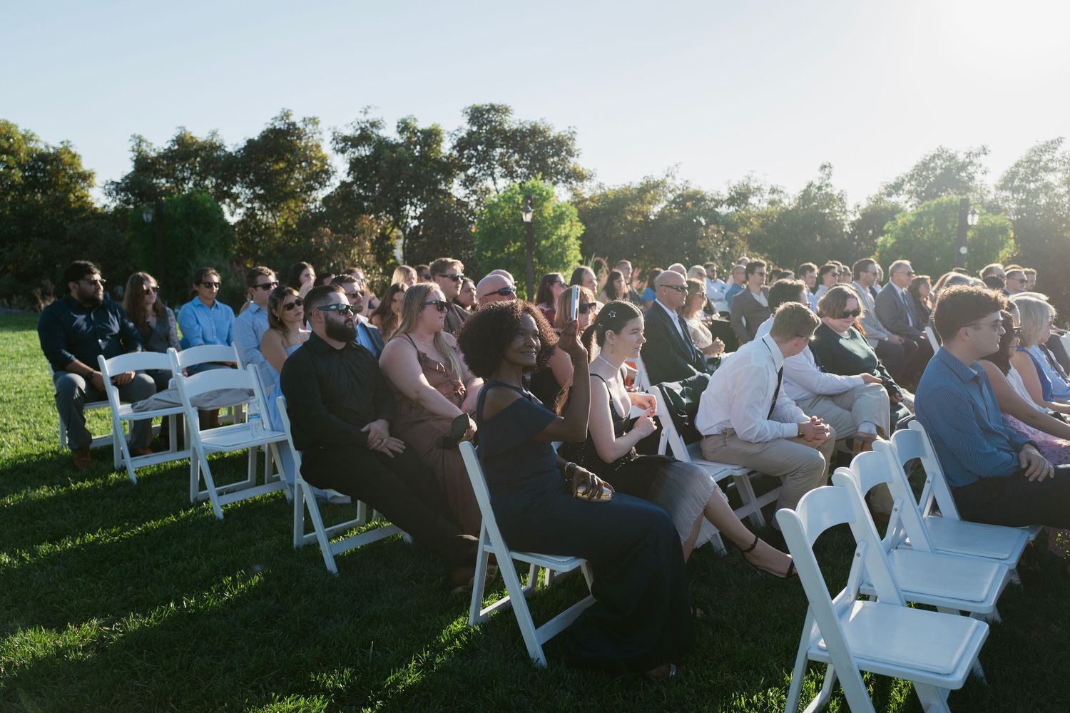 Large crowd of wedding guests seated in white chairs on green lawn during outdoor ceremony.
