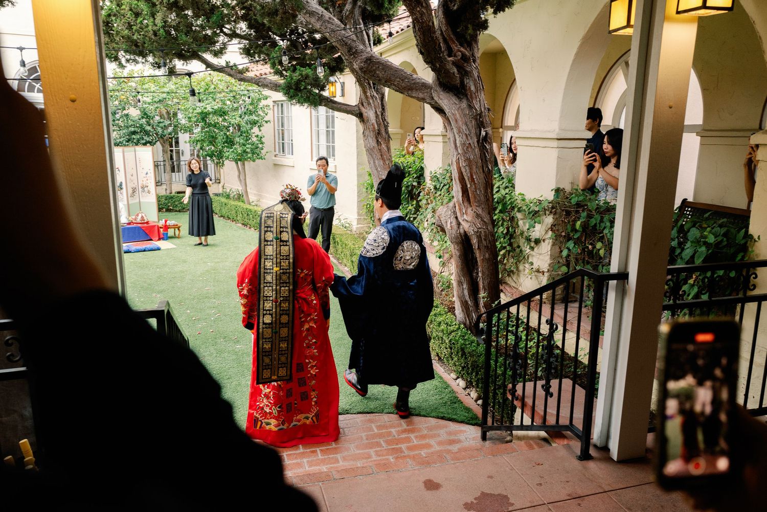 Couple in traditional Korean hanbok wedding attire walking through elegant garden courtyard.