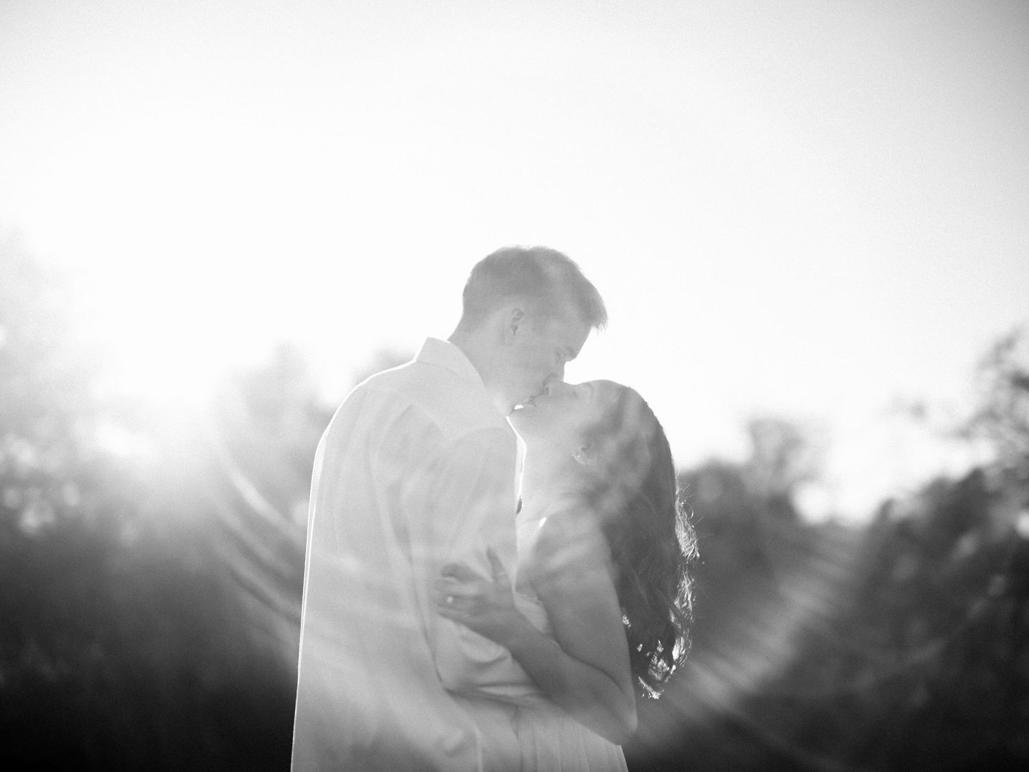 Black and white romantic couple embracing in sunlight, silhouetted against bright lens flare outdoors.