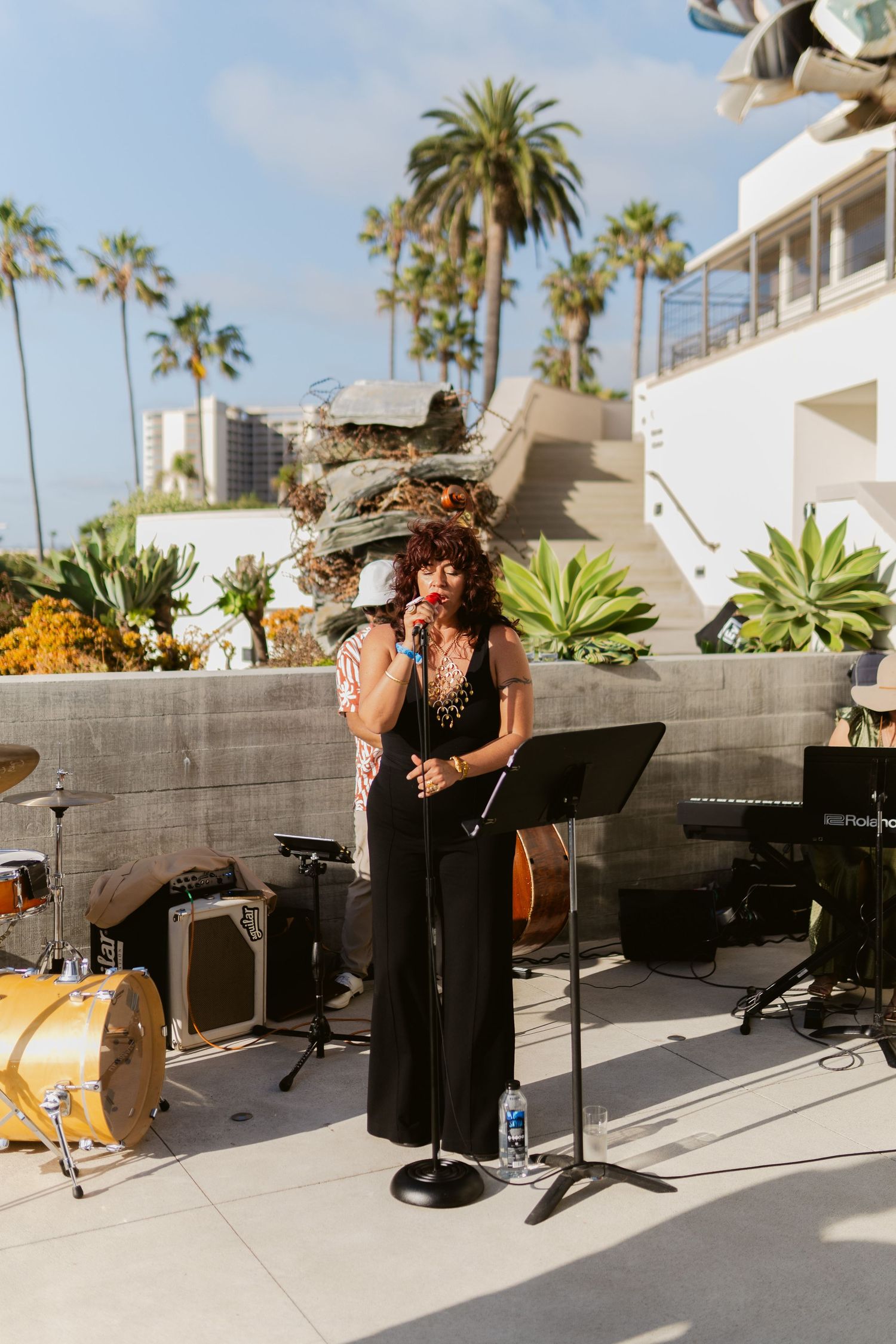 Two musicians performing outdoors at a sunny venue with palm trees, one singing into a microphone on a stand.