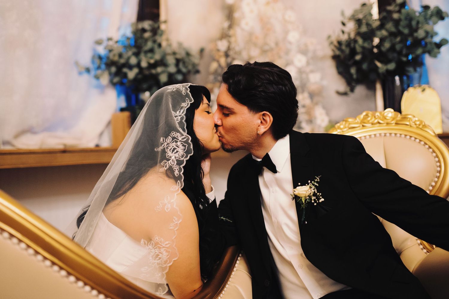 Bride and groom share a tender kiss while seated on a gold ornate sofa at reception.
