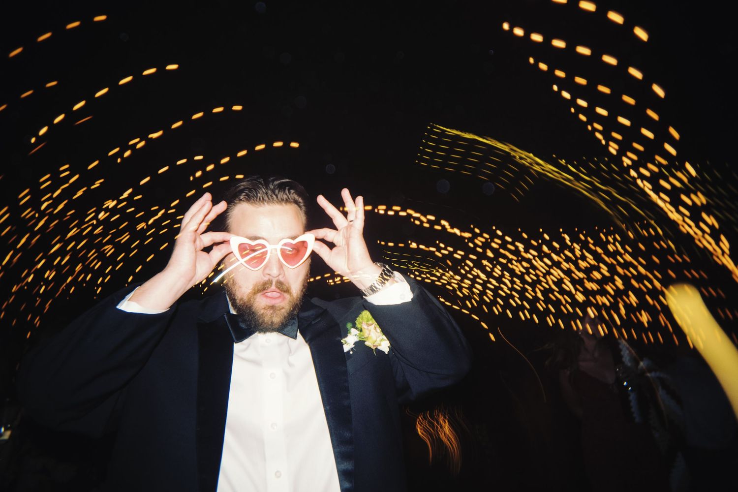 Groom in tuxedo wearing heart-shaped glasses posing at a dazzling nighttime wedding reception.