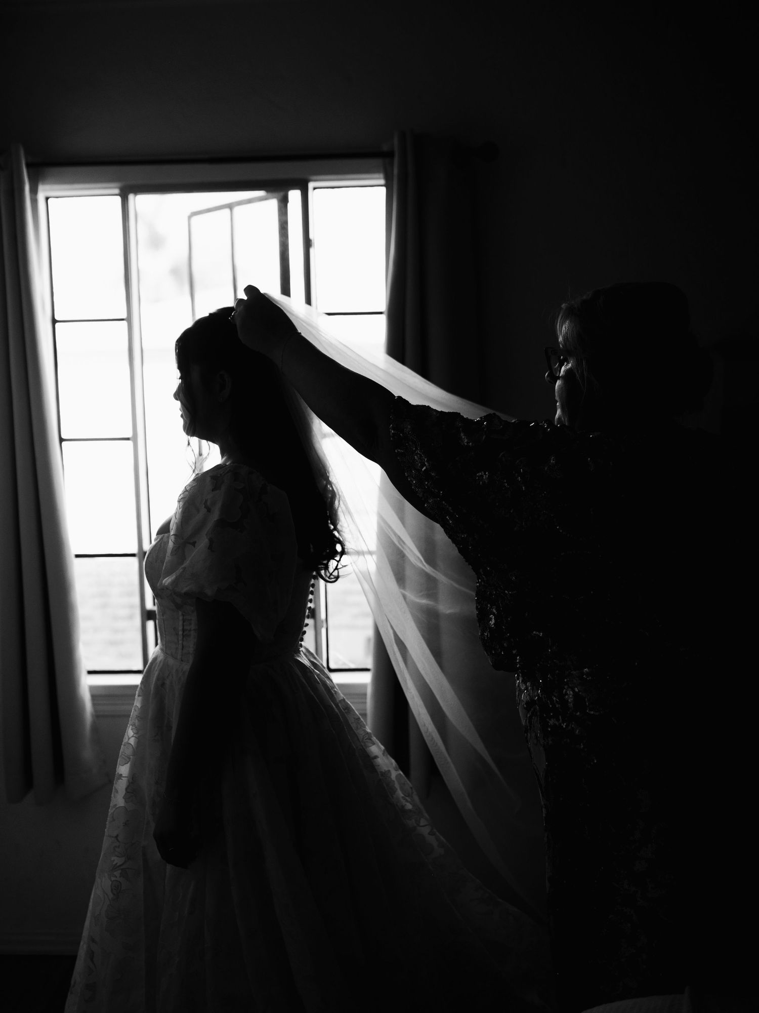 Black and white silhouette of bride having veil placed by helper in front of window light.
