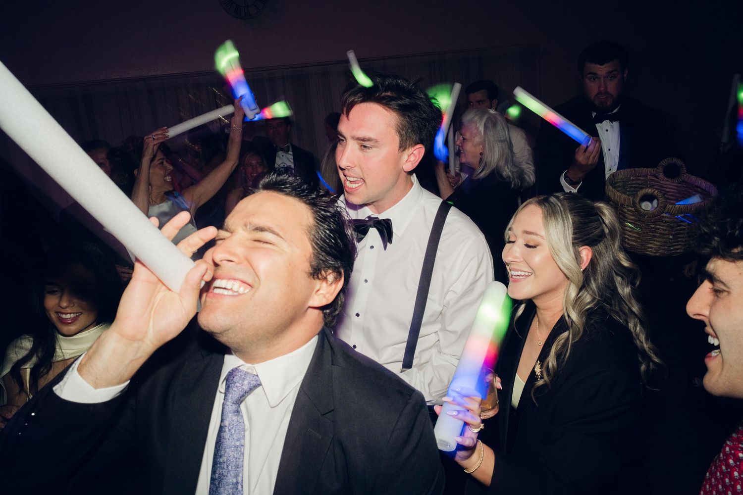 Guests celebrate at a lively wedding reception, holding glowing foam sticks and dancing under dim lights.