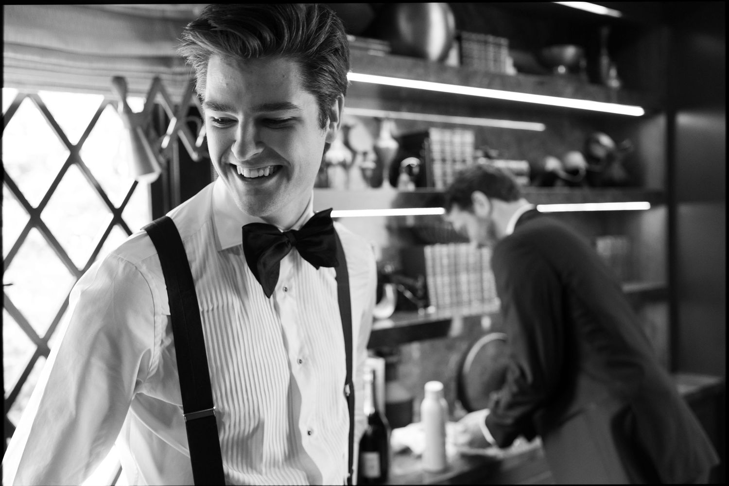 Black and white photo of young man in white shirt with suspenders and bow tie smiling in kitchen setting.