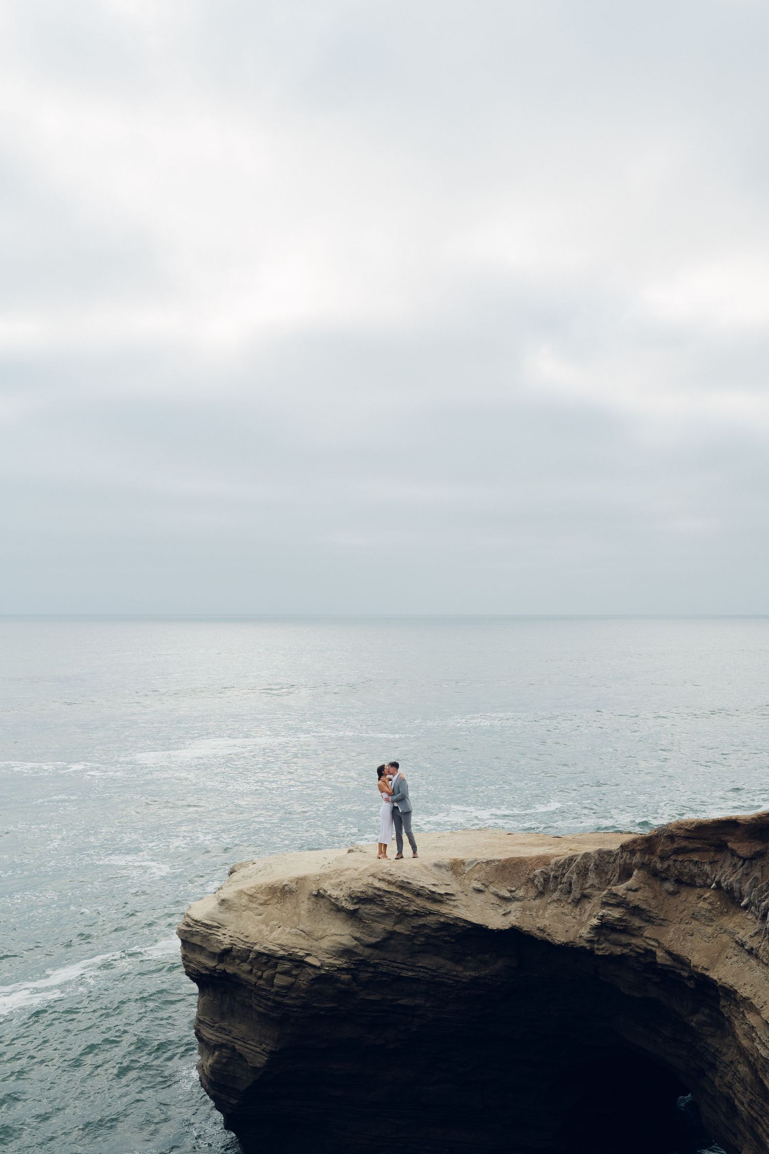 Couple embracing on a dramatic rocky cliff overlooking a vast, moody ocean landscape.
