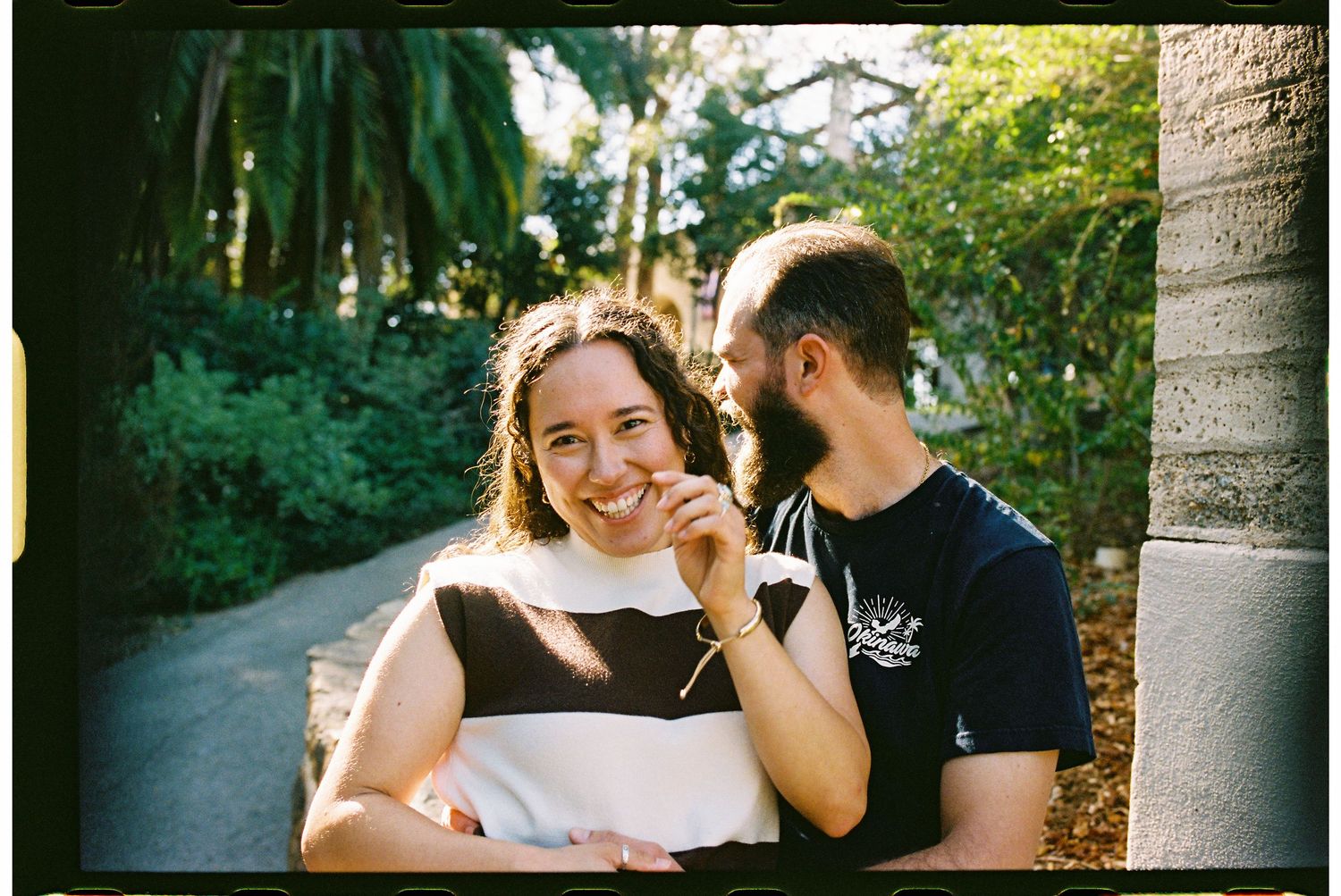 A smiling couple poses outdoors near a palm tree, surrounded by lush green foliage on a sunny day.