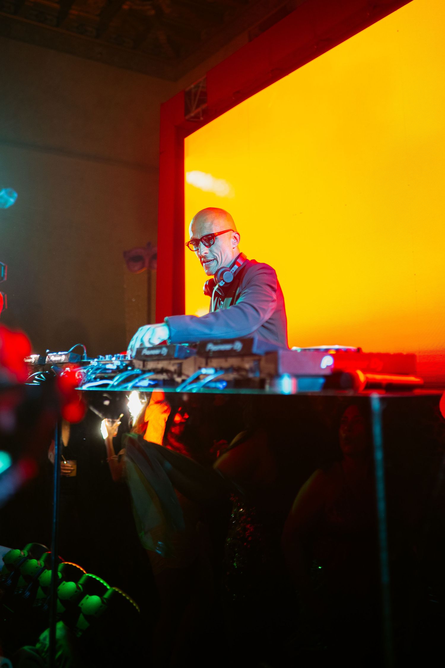 Bald DJ with sunglasses performing at a nightclub behind Pioneer DJ equipment under vibrant orange and red stage lighting.