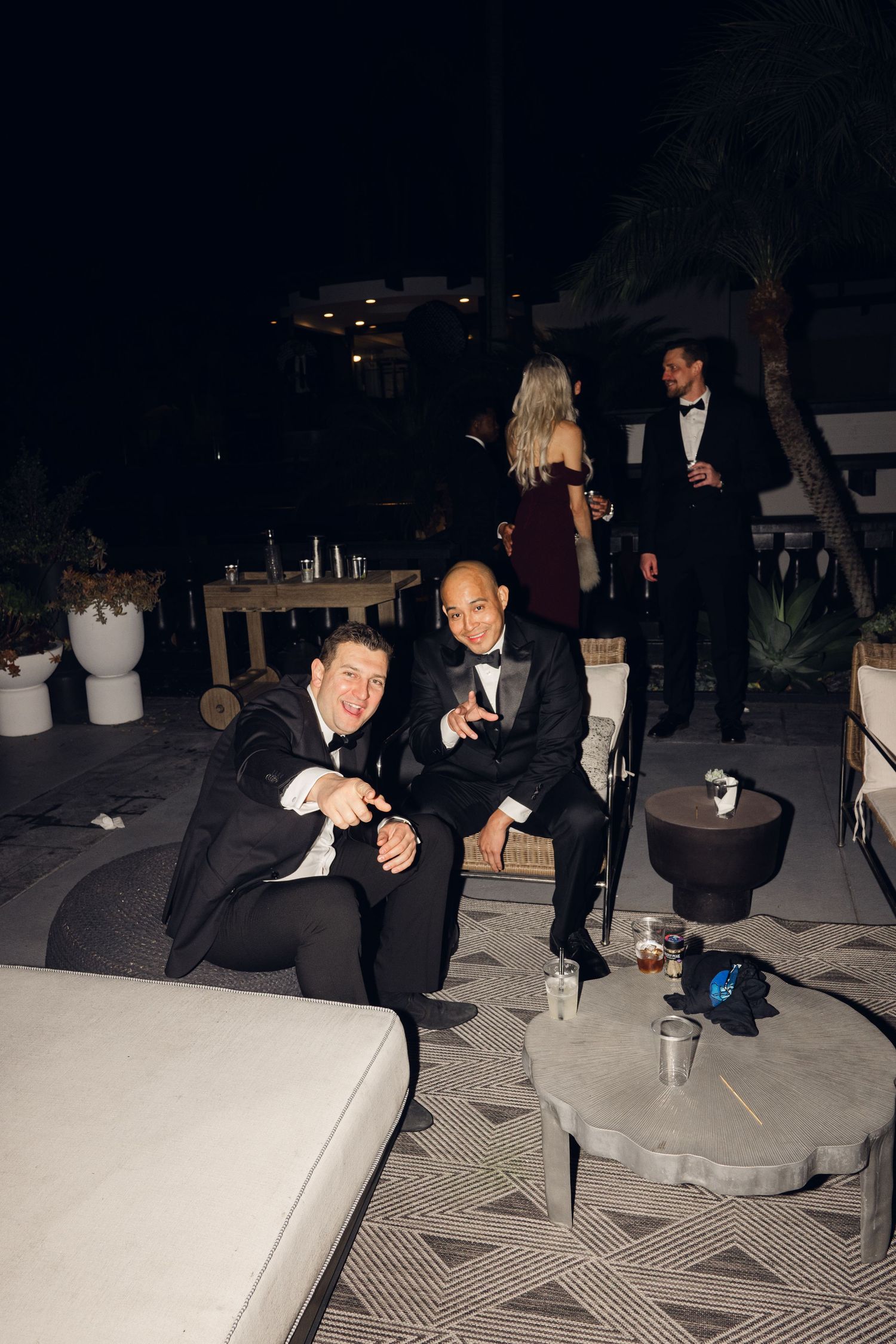 Two men in tuxedos pose and smile at a formal evening outdoor party with guests mingling in the background.