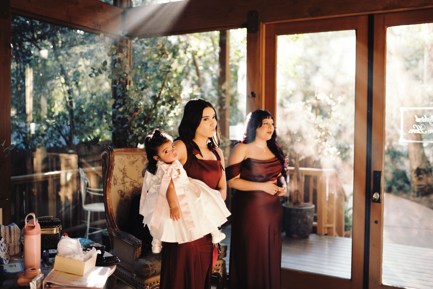Bridesmaids in burgundy dresses gather indoors, one holding a baby in a white dress.