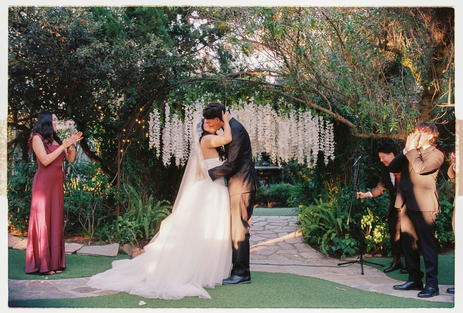 Bride and groom share first kiss under floral arch with bridesmaids and groomsmen celebrating beside them.