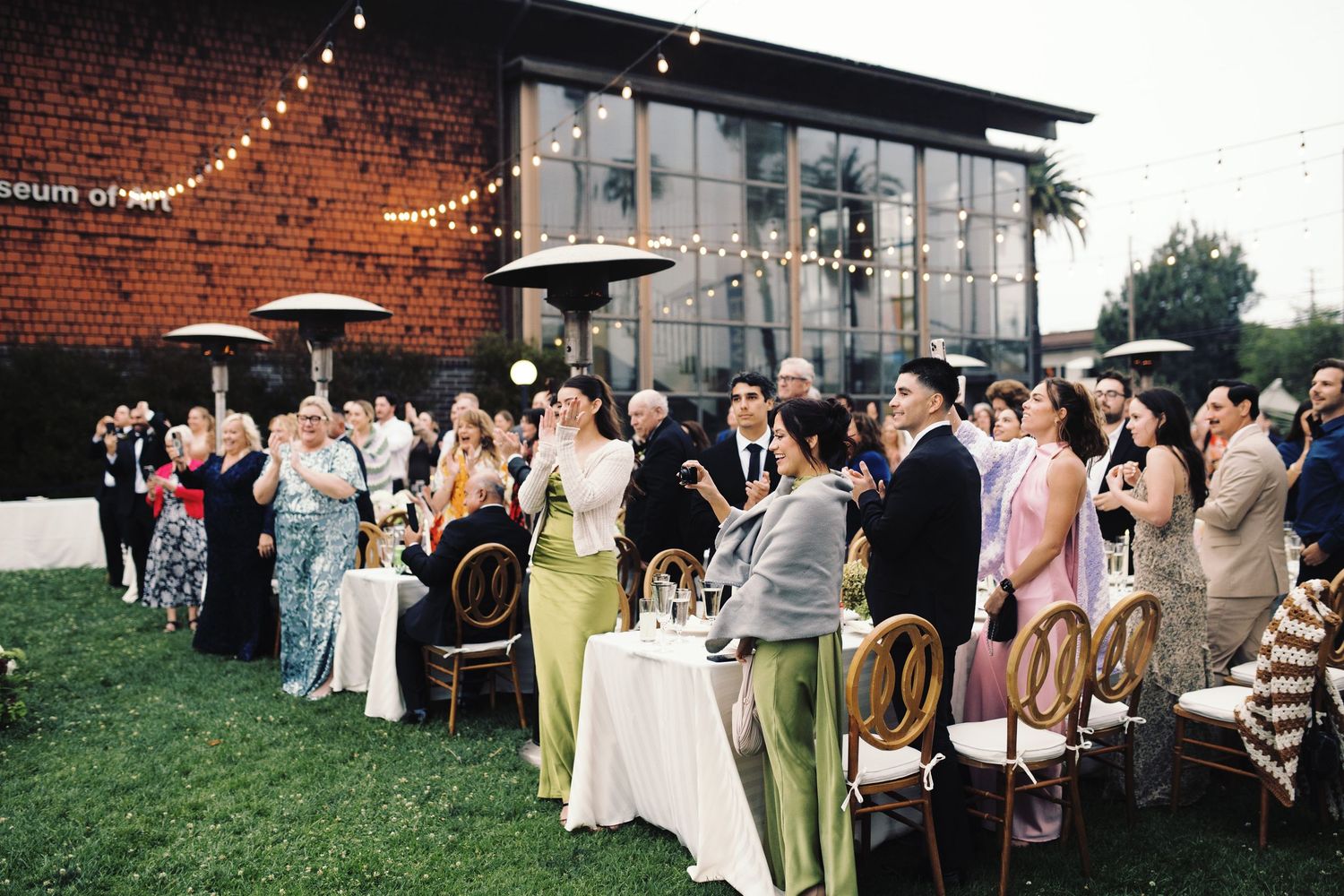 Outdoor daytime wedding reception with guests seated at tables under string lights at a museum venue.