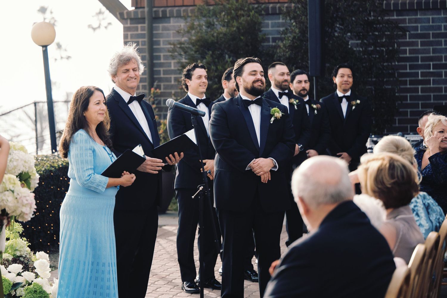 Groom in tuxedo smiles at altar during outdoor wedding ceremony with groomsmen and officiant.