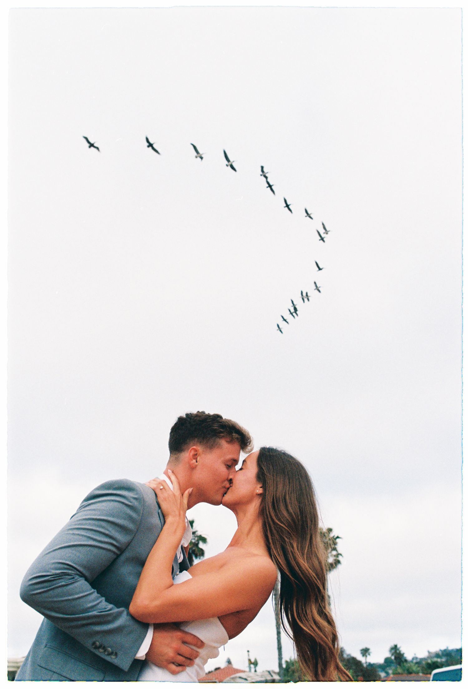 Newlyweds kissing outdoors with a flock of birds flying in a heart-like formation in the overcast sky above.