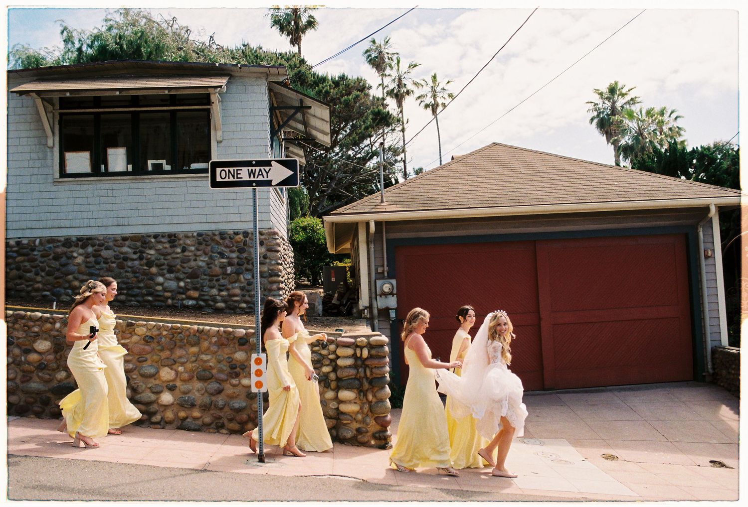 Bridesmaids in yellow gowns and bride in white walk together down a sunny residential street.