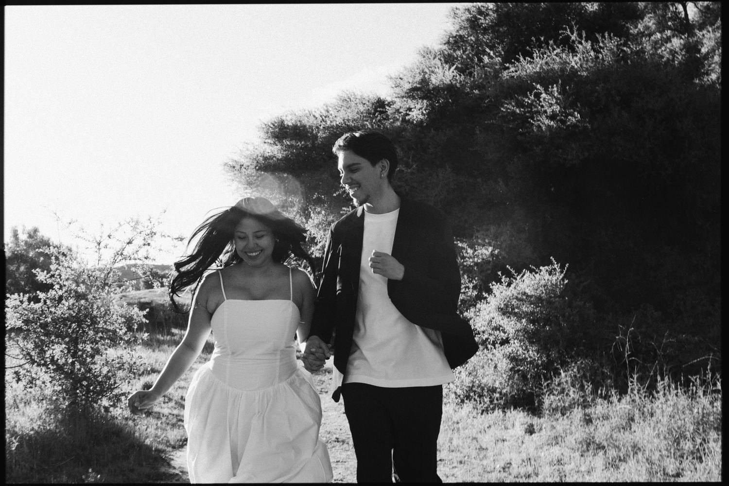 Black and white photo of a joyful couple running hand in hand through a sunny outdoor field.