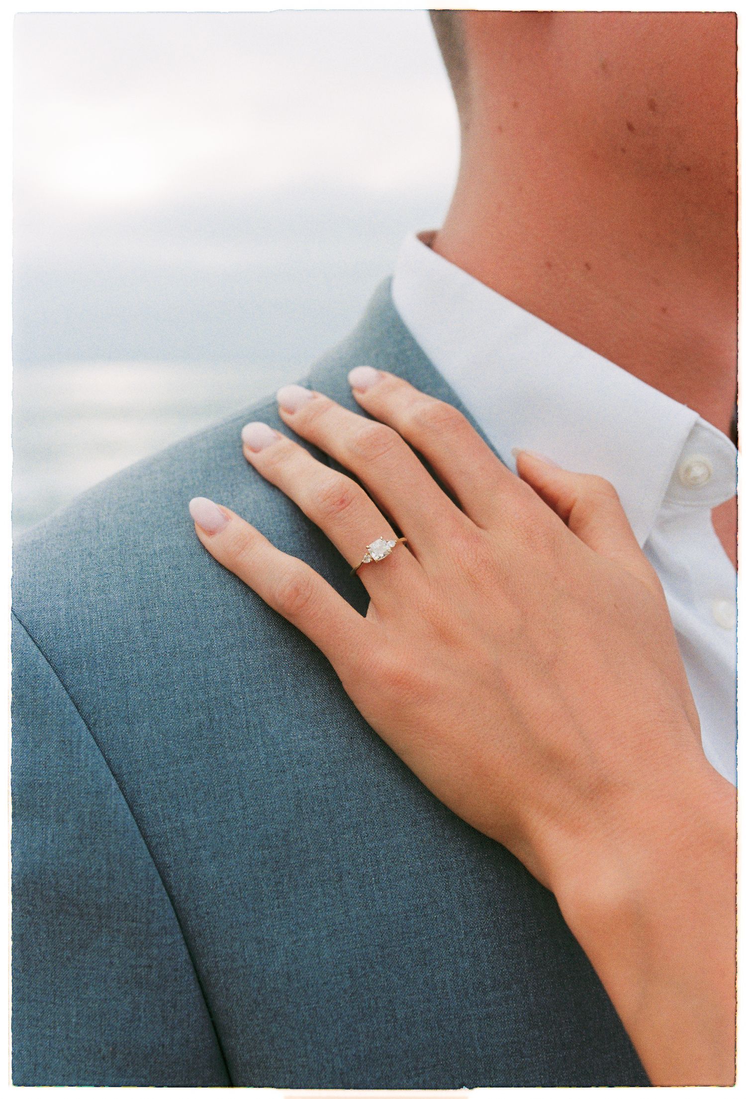 Close-up of elegant emerald-cut diamond engagement ring on bride's hand resting on groom's gray suit jacket shoulder.
