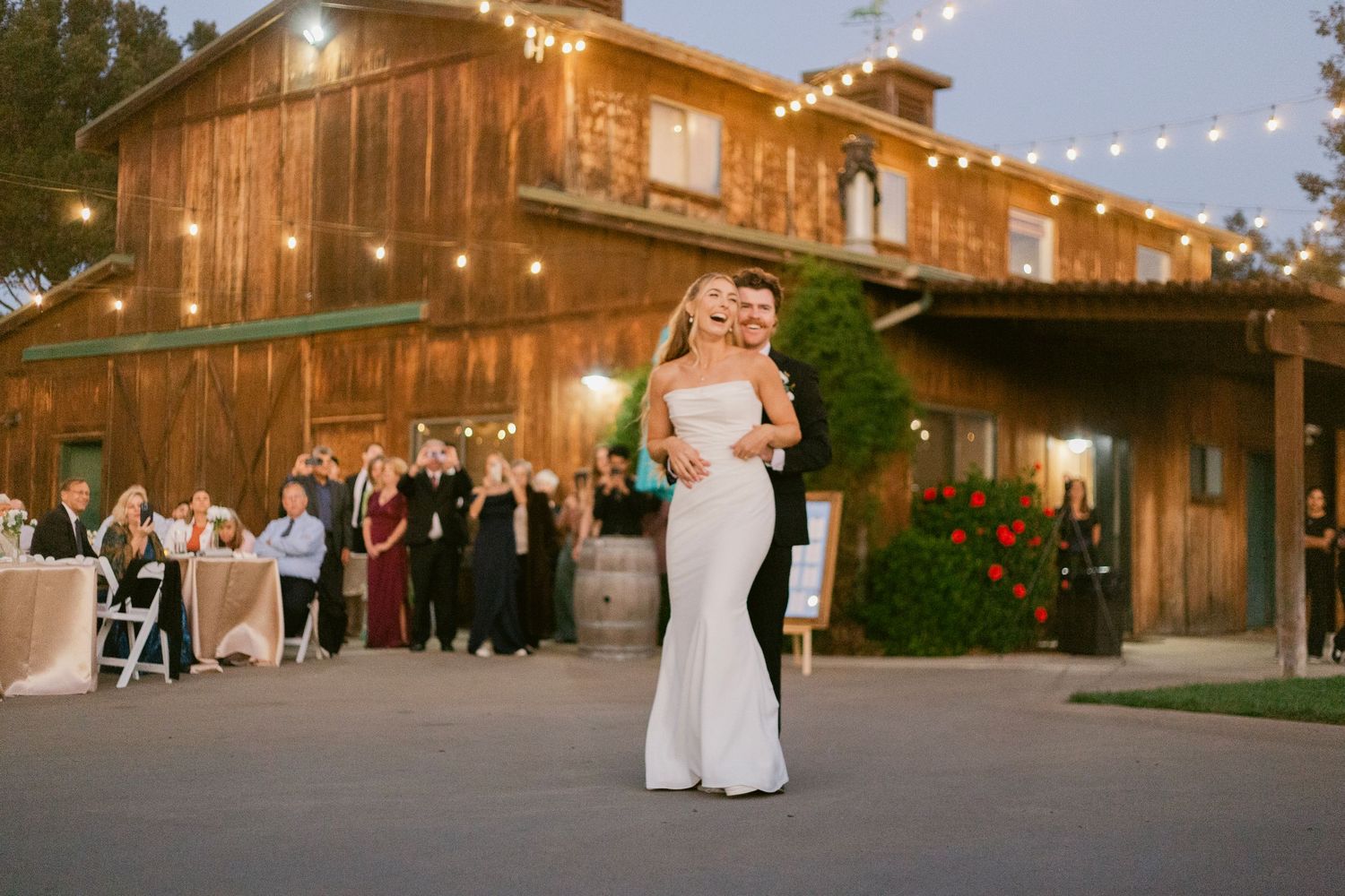 Bride and groom share first dance outside rustic barn wedding venue with string lights and guests watching.